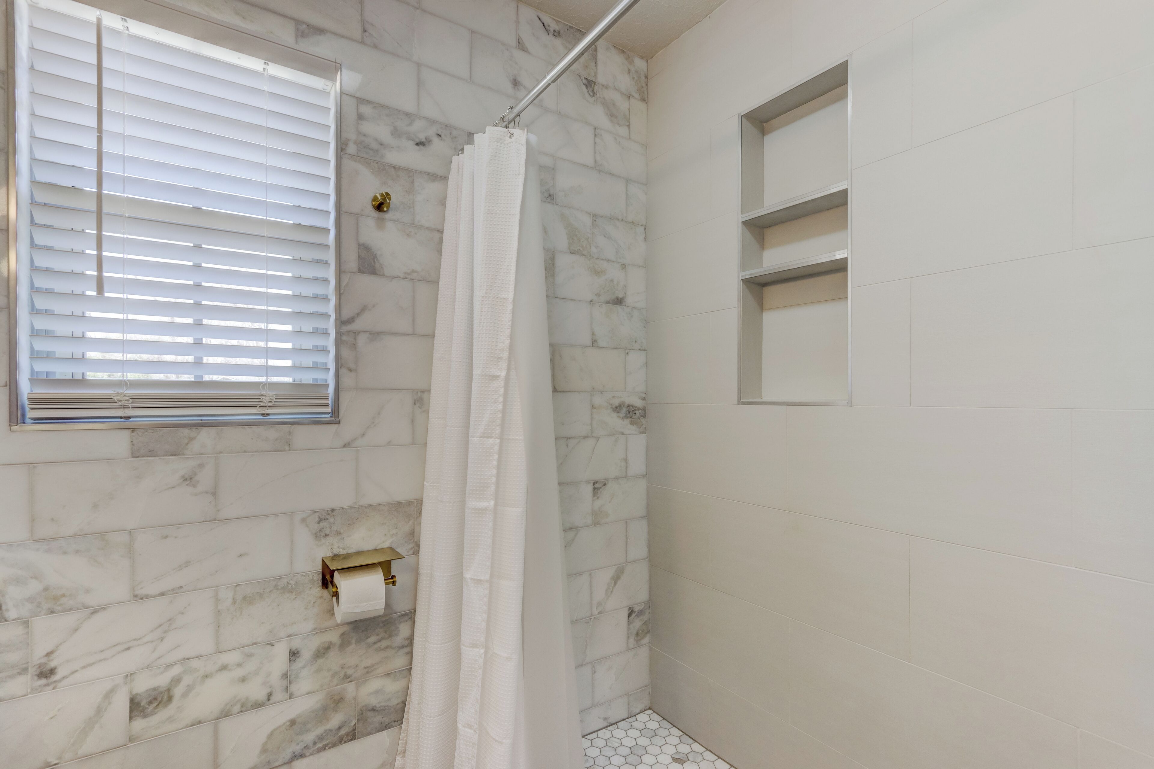 Un baño con baldosas de mármol, una cortina de ducha, una ventana con persianas y un dispensador de papel higiénico.