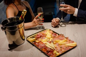 Un safata amb embutits i formatge damunt una taula, una ampolla de xampany en un cubell metàl·lic, i dues persones agafant copes de vi.