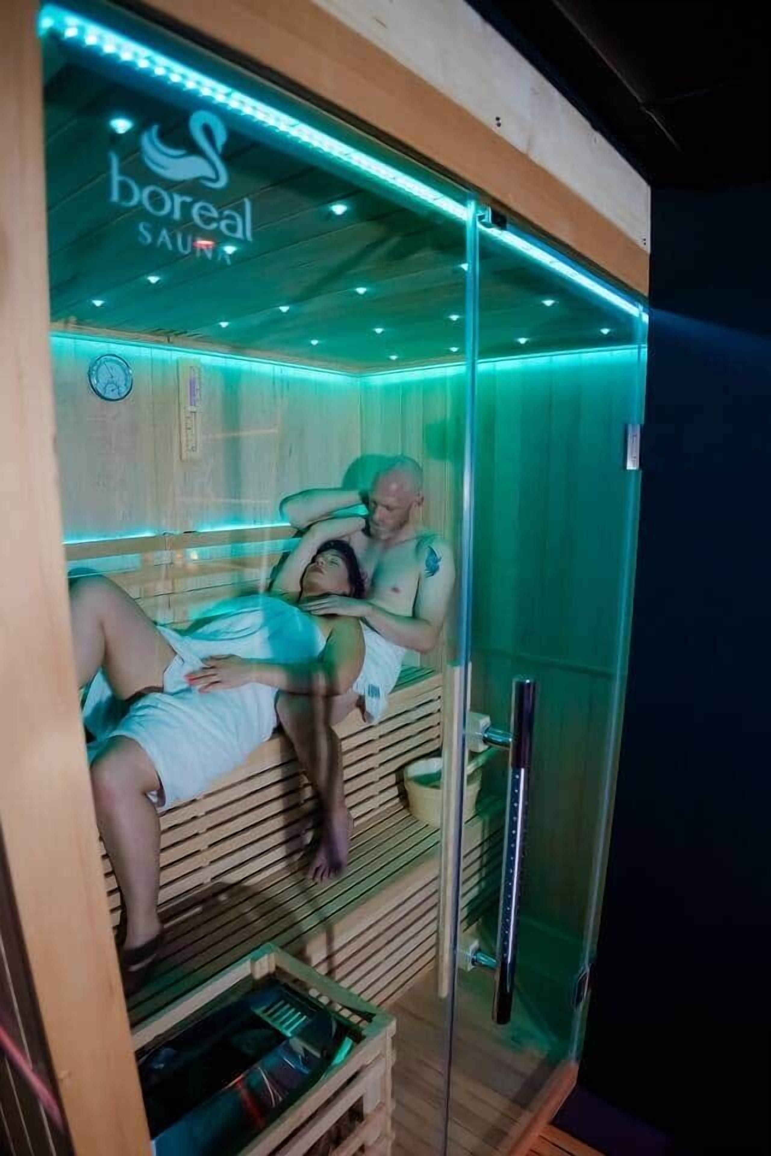 Een sauna met twee personen die op aparte banken liggen, een houten interieur en een glazen omheining.