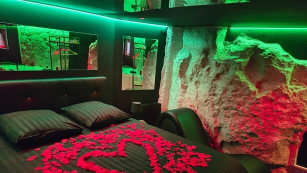 A bedroom with a bed covered in rose petals arranged in a heart shape, a large mirror, and a wall with a textured, rocky appearance. - La Cachette Secrete - Love Room Caen (Caen)