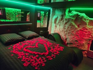 A bedroom with a bed covered in rose petals arranged in a heart shape, a large mirror, and a wall with a textured, rocky appearance. - La Cachette Secrete – Love Room in Caen (Caen)