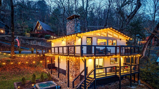A two-story cabin with a stone chimney, a hot tub, and a deck with string lights.