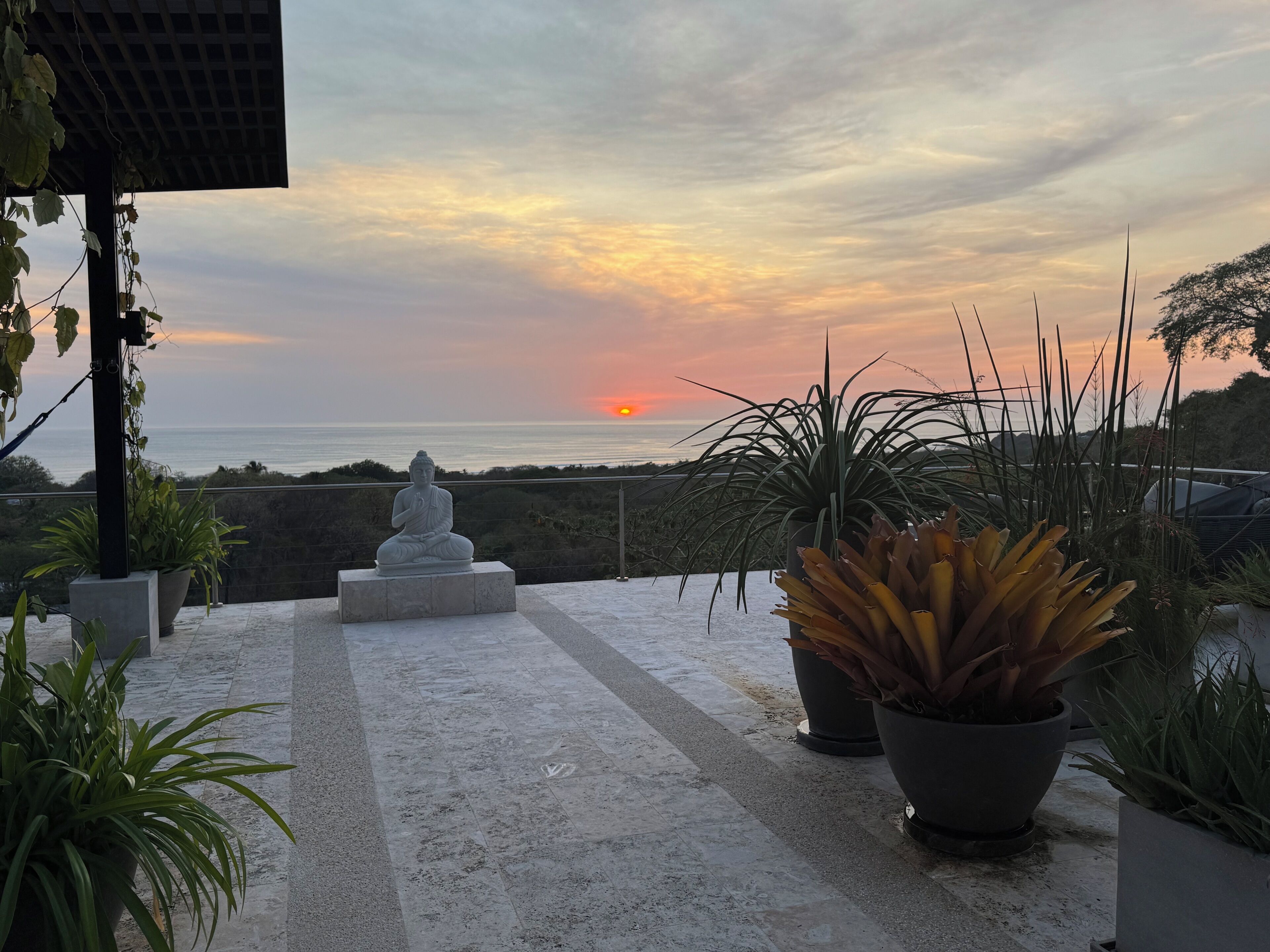 Un espace extérieur paisible, agrémenté d’une statue de Bouddha, de plantes en pot et offrant une vue sur le coucher de soleil sur l’océan.