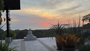 A serene outdoor area with a Buddha statue, potted plants, and a view of the sunset over the ocean.