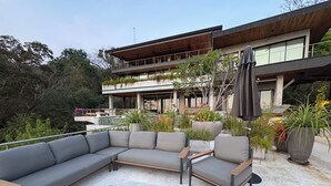 A modern outdoor seating area with a sectional sofa and chairs, surrounded by potted plants and a pergola.