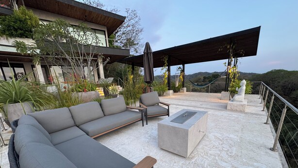 An outdoor seating area with a sofa, chairs, and a table, surrounded by plants and a view of the landscape.