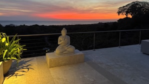 A statue on a balcony with a view of the sunset over the ocean.