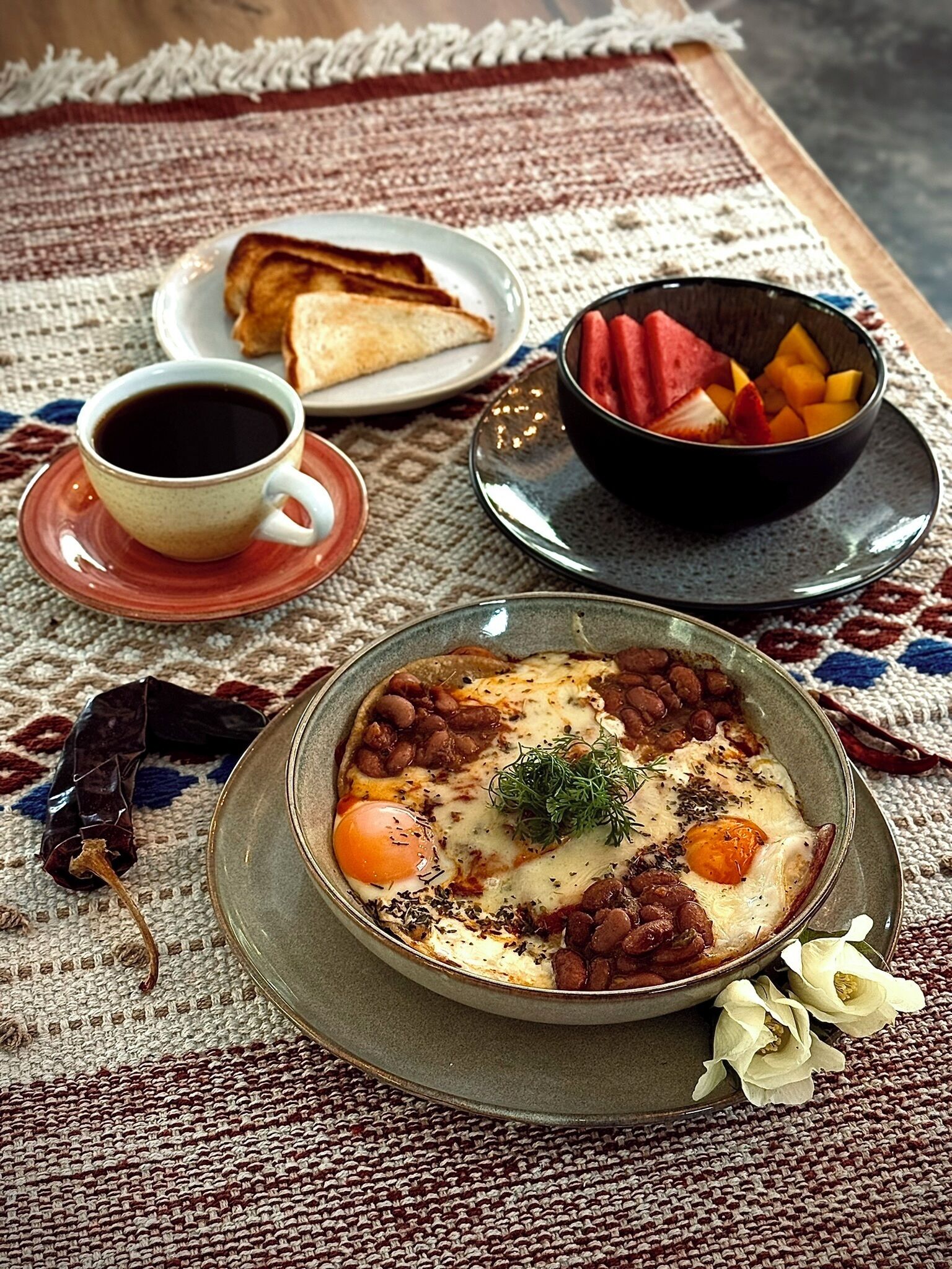 A breakfast setting with a bowl of beans and eggs, a cup of coffee, a plate of toast, and a bowl of fruit.