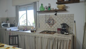 Une cuisine équipée d’un four à micro-ondes, d’un évier et d’une cuisinière. On y trouve une fenêtre avec des rideaux, une étagère avec des ustensiles de cuisine et une chaise.