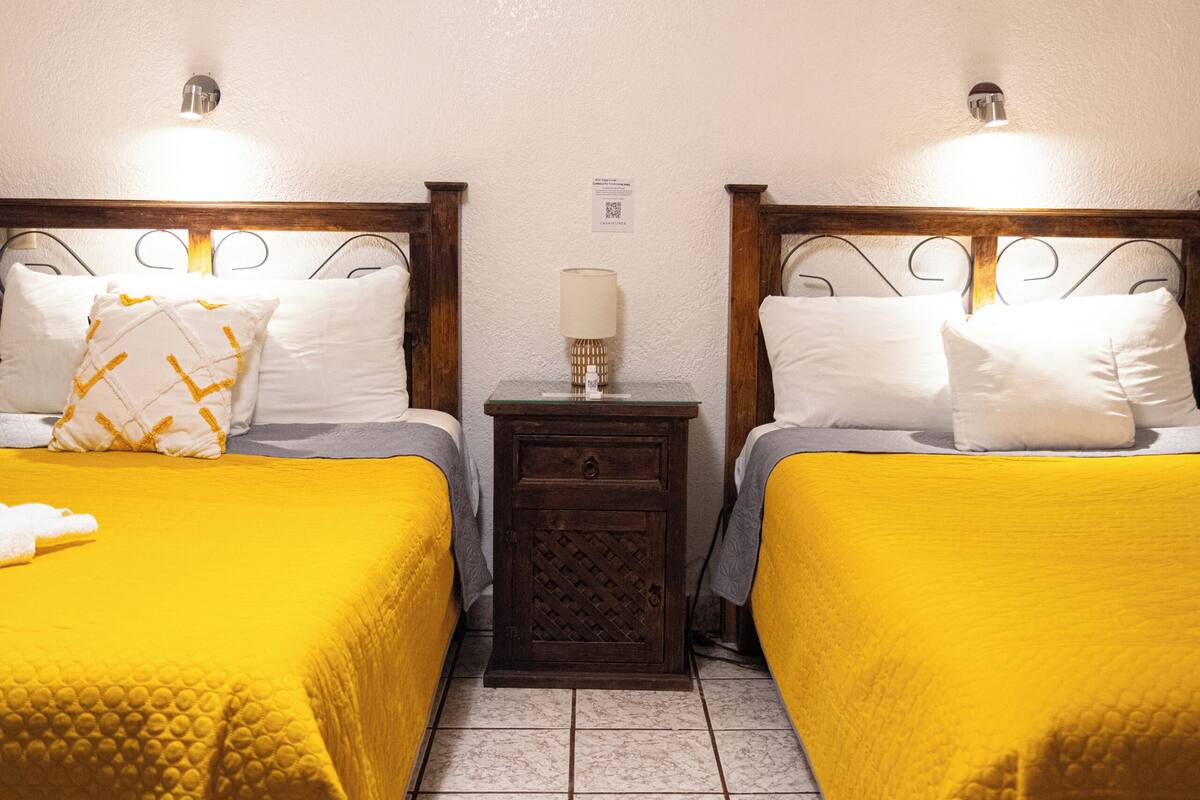A hotel room with two beds, each with a yellow bedspread and white pillows, a nightstand with a lamp, and a tiled floor.