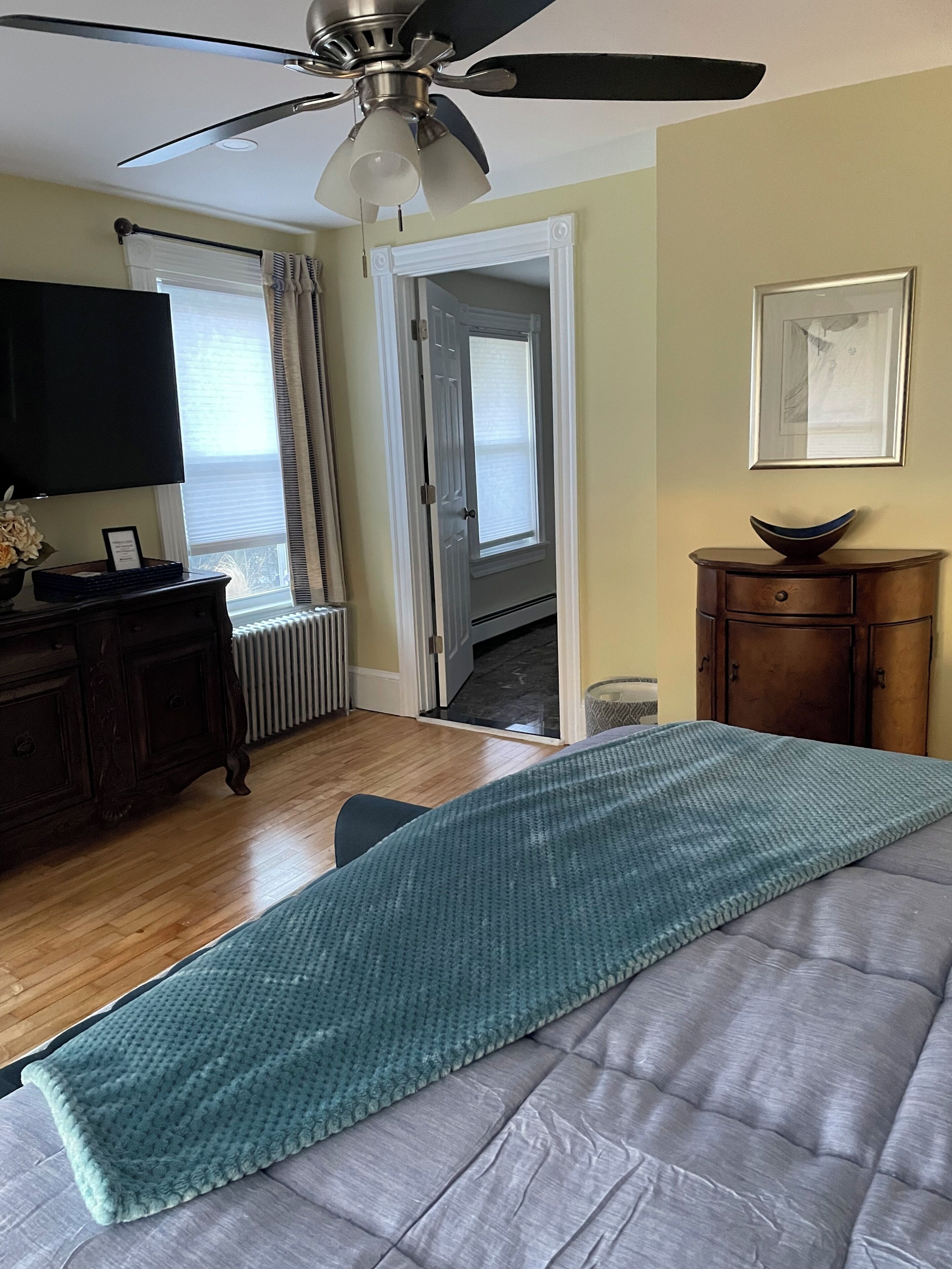 Une chambre à coucher comprenant un lit, un ventilateur de plafond, une commode, un téléviseur et une porte donnant sur une autre pièce.