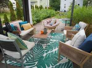 Un espace extérieur aménagé avec des meubles en osier, un tapis à motifs et une vue sur un jardin et des bâtiments en arrière-plan.