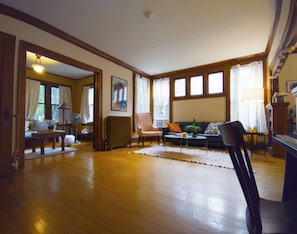 A spacious living room with hardwood flooring, a sofa, a coffee table, and a piano. There are windows with curtains, a painting on the wall, and a door leading to another room.