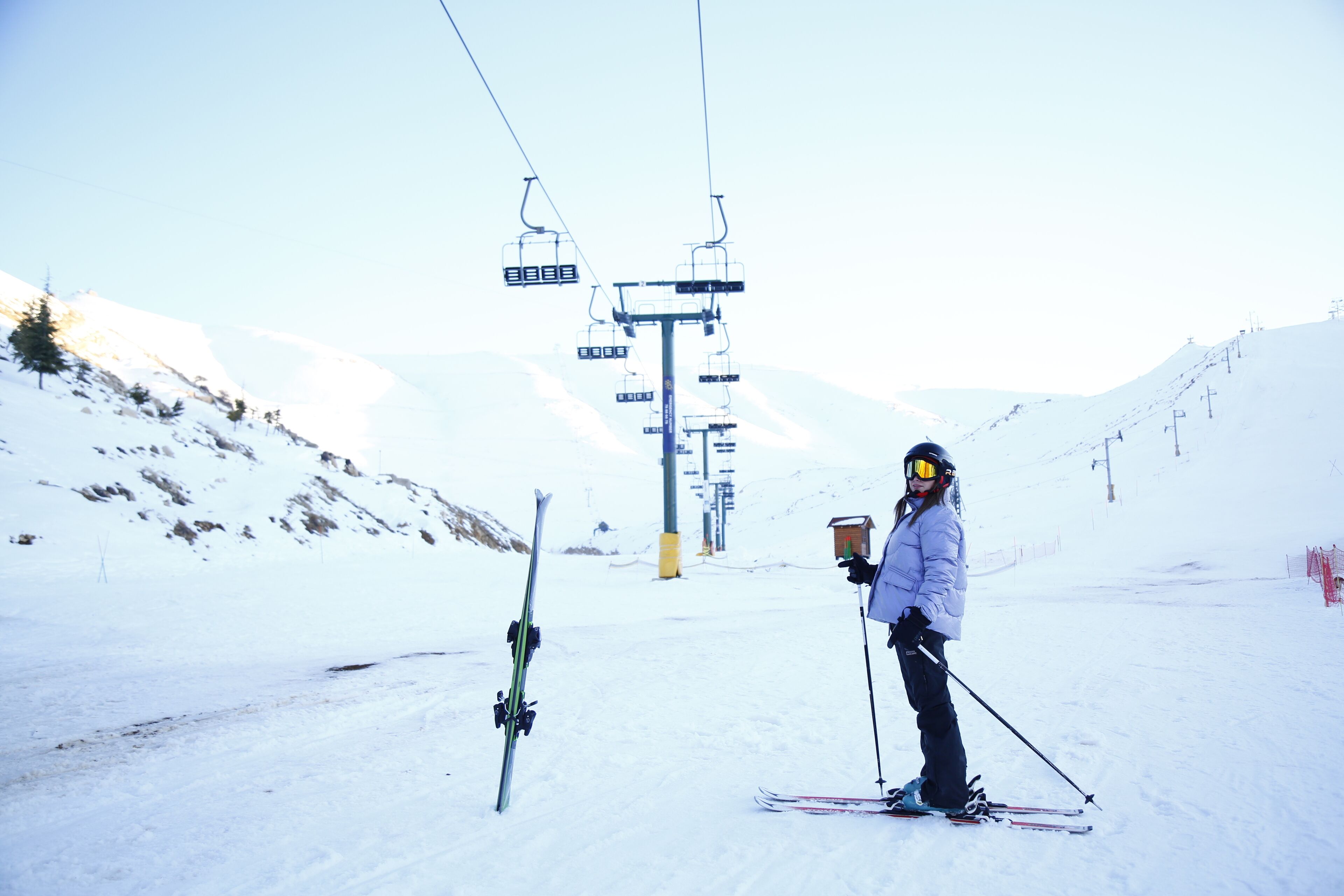 A person standing on a snowy ski slope with ski equipment, a ski lift in the background, and a clear sky.