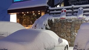 Lumet peittänyt auto pysäköitynä rakennuksen eteen, ja rakennuksen kyltissä lukee: ”Itävallan vuoristomajat, moottorikelkat”.