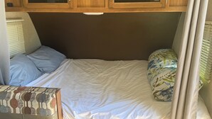A compact RV bedroom with a bed, a small table, and wooden cabinets with glass doors. - Cozy Vegas Bunkhouse RV (Las Vegas)
