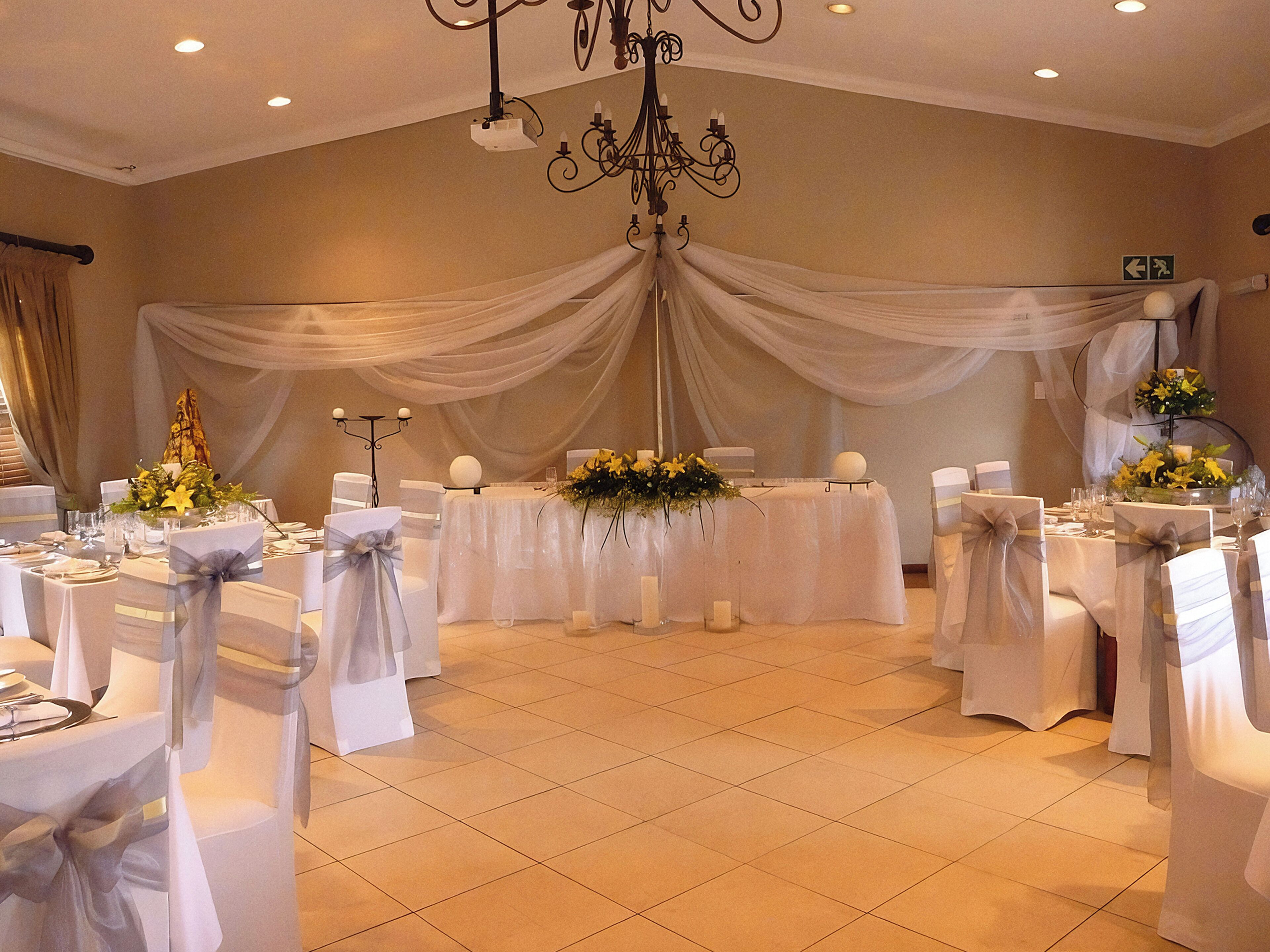 A banquet hall with white draped curtains, a chandelier, and tables set for a formal event.