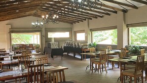 Ein Restaurant mit Holztischen und -stühlen, einer Kristallleuchter und großen Fenstern mit Grünpflanzen davor.