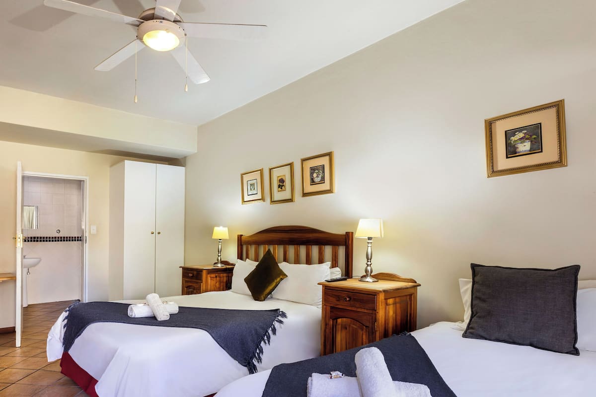 Une chambre d’hôtel avec deux lits, un ventilateur de plafond, des lampes de chevet, une tête de lit en bois, une table de chevet, un tableau encadré au mur et une salle de bain avec un lavabo et un miroir.