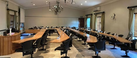 Uma sala de conferências com uma mesa comprida, cadeiras, um lustre e janelas com cortinas.