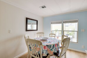 Ein Essbereich mit einem Tisch, der mit einer blumigen Tischdecke bedeckt ist, umgeben von vier weißen Stühlen. Rechts befindet sich ein Fenster mit Jalousien, und über dem Tisch ist ein wandmontierter Spiegel angebracht.