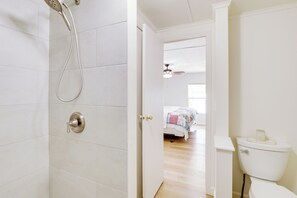 Ein Badezimmer mit Dusche, Toilette und einer Tür zu einem Schlafzimmer mit Bett und Deckenventilator.