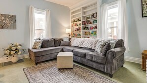 A living room with a sectional sofa, a side table, a bookshelf, and a window with curtains. - Walkable Group Getaway w/ Bikes in West Liberty! (West Liberty)