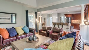 Un salón espacioso con un amplio sofá de cuero marrón, una mesa de centro de madera y cojines decorativos de colores. Se aprecia una zona de comedor con cocina al fondo.