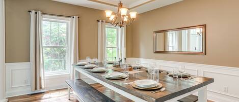 Une salle à manger avec une longue table dressée pour six personnes, un lustre et de grandes fenêtres ornées de rideaux.
