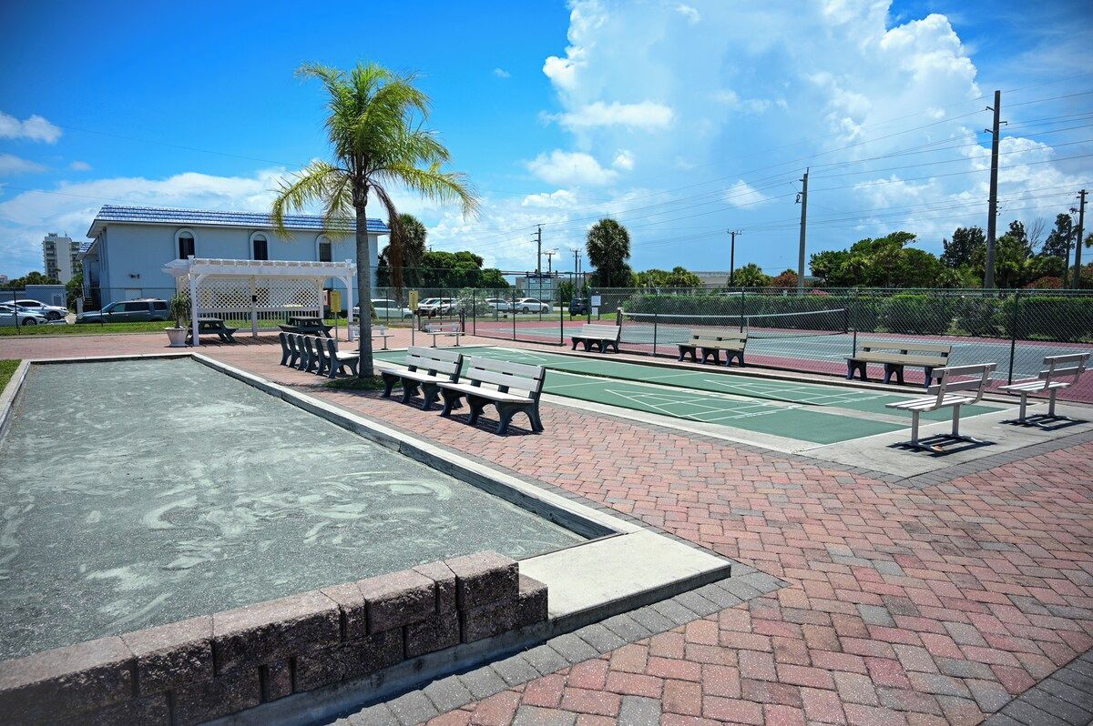 A paved area with benches and a basketball court, surrounded by a fence and palm trees.