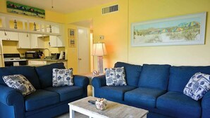 A living room with two blue sofas, a coffee table, and a kitchen in the background. - Direct Ocean Complex! Diplomat 220-21 (Cocoa Beach)