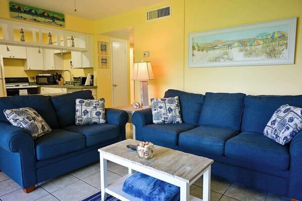 A living room with two blue sofas, a coffee table, and a kitchen in the background.