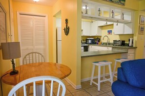 A compact kitchen with white cabinets, a dining area with a wooden table and chairs, and a blue sofa. - 2BR Direct Ocean Complex • Beach Access • Pool • Free Parking (Cocoa Beach)
