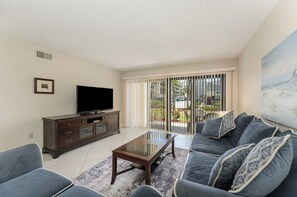 A living room with a flat-screen TV, dark-colored sofa, and a glass coffee table. - Steps to the Ocean • 2BR Condo • Pool (Cocoa Beach)