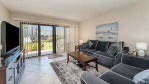 A modern living room with a sofa, coffee table, TV, and a painting of a lighthouse. - Steps to Sand! Ground-Floor 2BR by Pool (Cocoa Beach)
