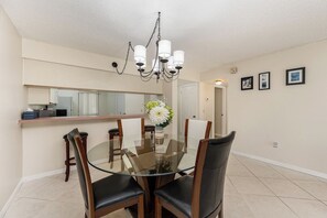 A dining area with a glass table, wooden chairs, a chandelier, and a kitchen in the background.