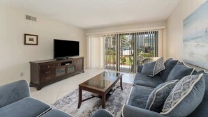 A living room with a sofa set, a coffee table, a TV on a cabinet, and a sliding glass door leading to an outdoor area. - Steps to Sand! Ground-Floor 2BR by Pool (Cocoa Beach)