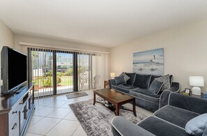 A modern living room with a sofa, coffee table, TV, and a painting of a lighthouse. - Steps to the Ocean • 2BR Condo • Pool (Cocoa Beach)