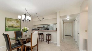 A dining area with a glass table, four chairs, a chandelier, a kitchen area with a microwave, and a hallway leading to a door. - Steps to Sand! Ground-Floor 2BR by Pool (Cocoa Beach)
