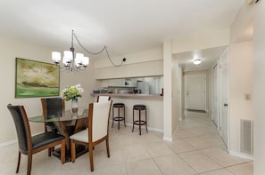 A dining area with a glass table, chairs, a chandelier, and a kitchen in the background. - Steps to the Ocean • 2BR Condo • Pool (Cocoa Beach)