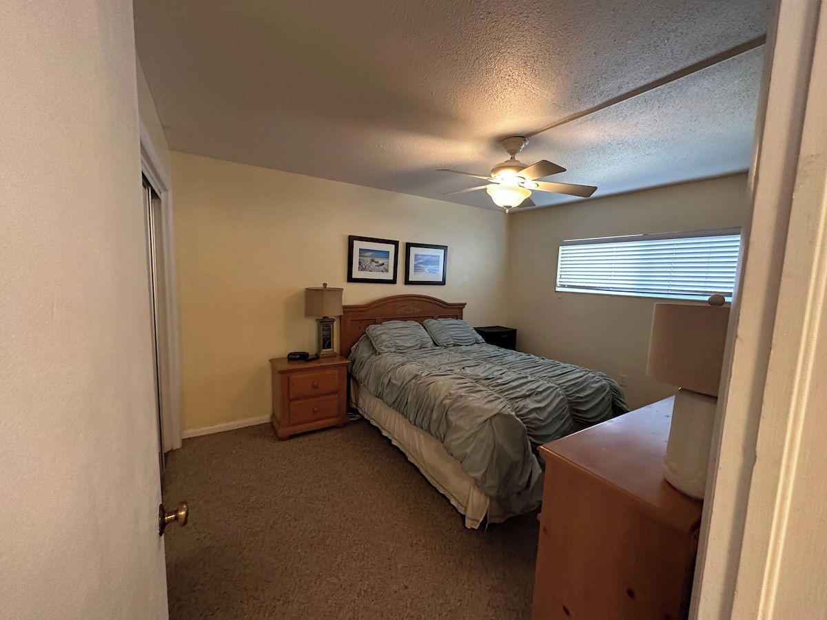 A bedroom with a bed, nightstands, a ceiling fan, a window with blinds, and a door.