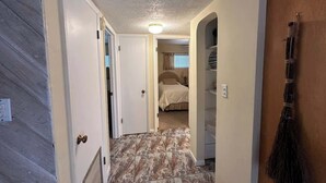 A narrow hallway with a bed in the background, a broom on the wall, and a door on the left.