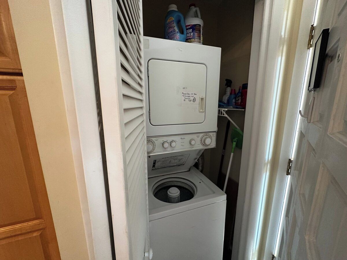 Une petite buanderie équipée d’un lave-linge et d’un sèche-linge à chargement frontal, d’une hotte aspirante et d’un étagère où sont rangés les produits de nettoyage.
