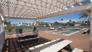 An outdoor tennis court with a covered seating area, picnic tables, and a trash can.