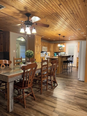 Une cuisine aux murs lambrés, avec un coin repas, dotée d’un ventilateur de plafond, de luminaires suspendus et d’un plancher en bois.