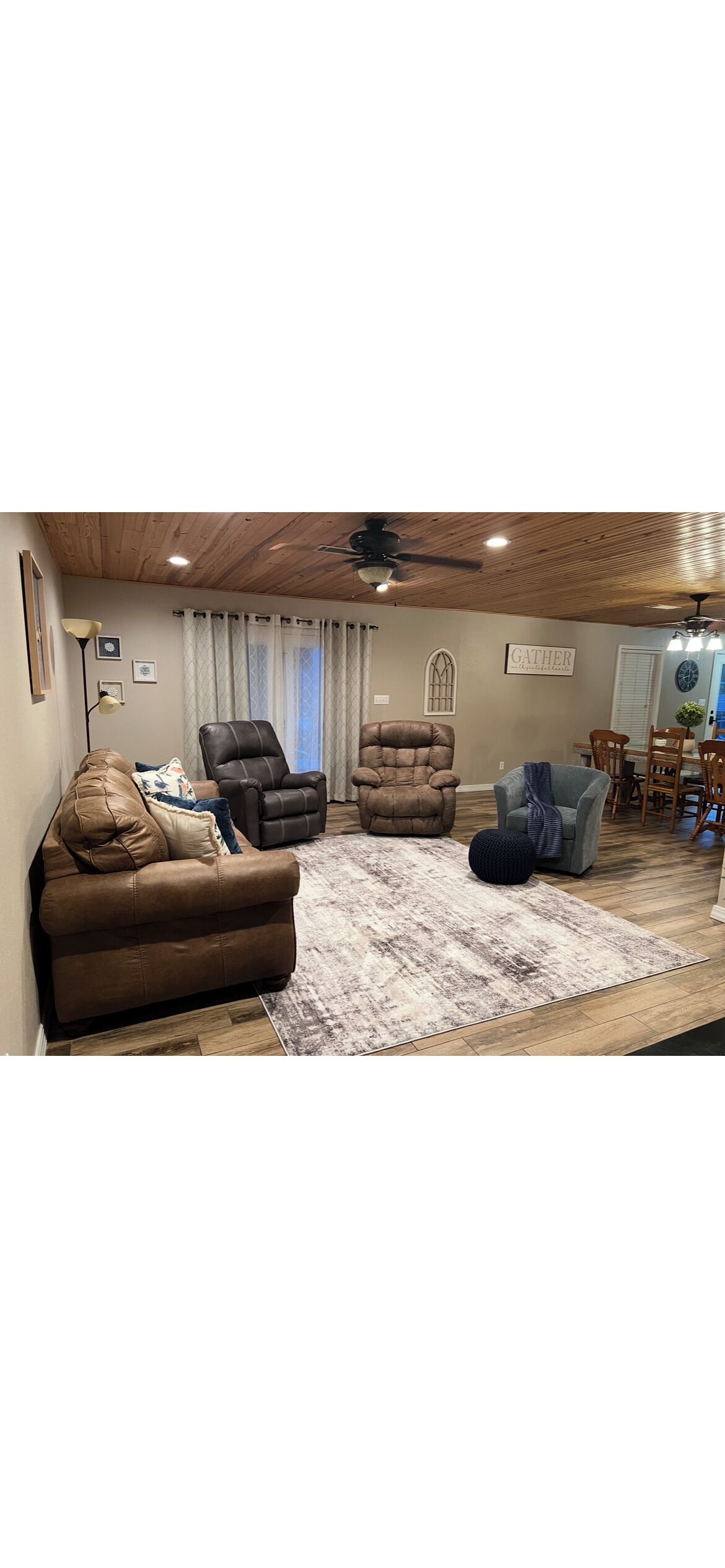 A cozy living room with a brown leather sofa, two armchairs, a blue ottoman, and a wooden ceiling.