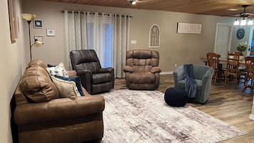 A cozy living room with a brown leather sofa, two armchairs, a blue ottoman, and a wooden ceiling.