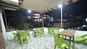 Un espace extérieur aménagé en terrasse, avec des chaises et des tables en plastique vertes, un sol carrelé et une vue sur les bâtiments en arrière-plan.