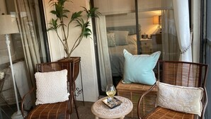 Un rincón acogedor con sillones de mimbre y una pequeña mesa redonda, una planta en maceta, y una cama con una mesita de noche al fondo.