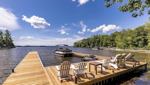 Une jetée en bois, agrémentée de chaises Adirondack et d’une table, s’avance sur un lac paisible où un bateau est amarré à proximité.